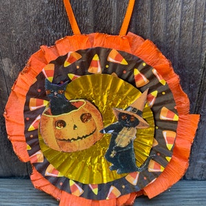 K&ouml;nnte beinhalten: Halloween-Dekoration mit einem mehrschichtigen kreisf&ouml;rmigen Design. Die Mitte hat eine goldene F&auml;cherform, umgeben von einer braunen Schicht mit S&uuml;&szlig;igkeiten-Mais-Mustern. Zwei Illustrationen von schwarzen Katzen mit Hexenh&uuml;ten und einem K&uuml;rbis sind abgebildet.