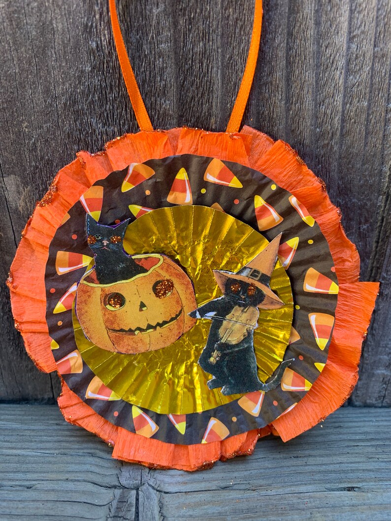 K&ouml;nnte beinhalten: Halloween-Dekoration mit einem mehrschichtigen kreisf&ouml;rmigen Design. Die Mitte hat eine goldene F&auml;cherform, umgeben von einer braunen Schicht mit S&uuml;&szlig;igkeiten-Mais-Mustern. Zwei Illustrationen von schwarzen Katzen mit Hexenh&uuml;ten und einem K&uuml;rbis sind abgebildet.