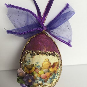 May include: Purple Easter egg ornament with a decorative floral and bird design. The egg is adorned with a gold glitter trim and a purple ribbon for hanging. A small cluster of flowers is attached to the egg.