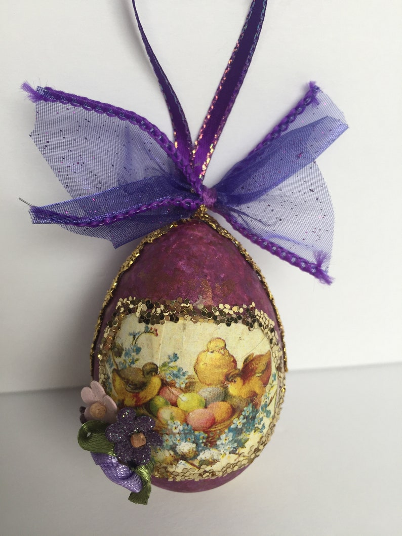 May include: Purple Easter egg ornament with a decorative floral and bird design. The egg is adorned with a gold glitter trim and a purple ribbon for hanging. A small cluster of flowers is attached to the egg.