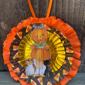 K&ouml;nnte beinhalten: Ein Halloween-Ornament mit einer K&uuml;rbiskopffigur, einer Eule und einer schwarzen Katze. Das Ornament hat Schichten aus orangefarbenem und goldenem Papier mit Zuckermais-Akzenten. Ein orangefarbenes Band ist zum Aufh&auml;ngen befestigt.