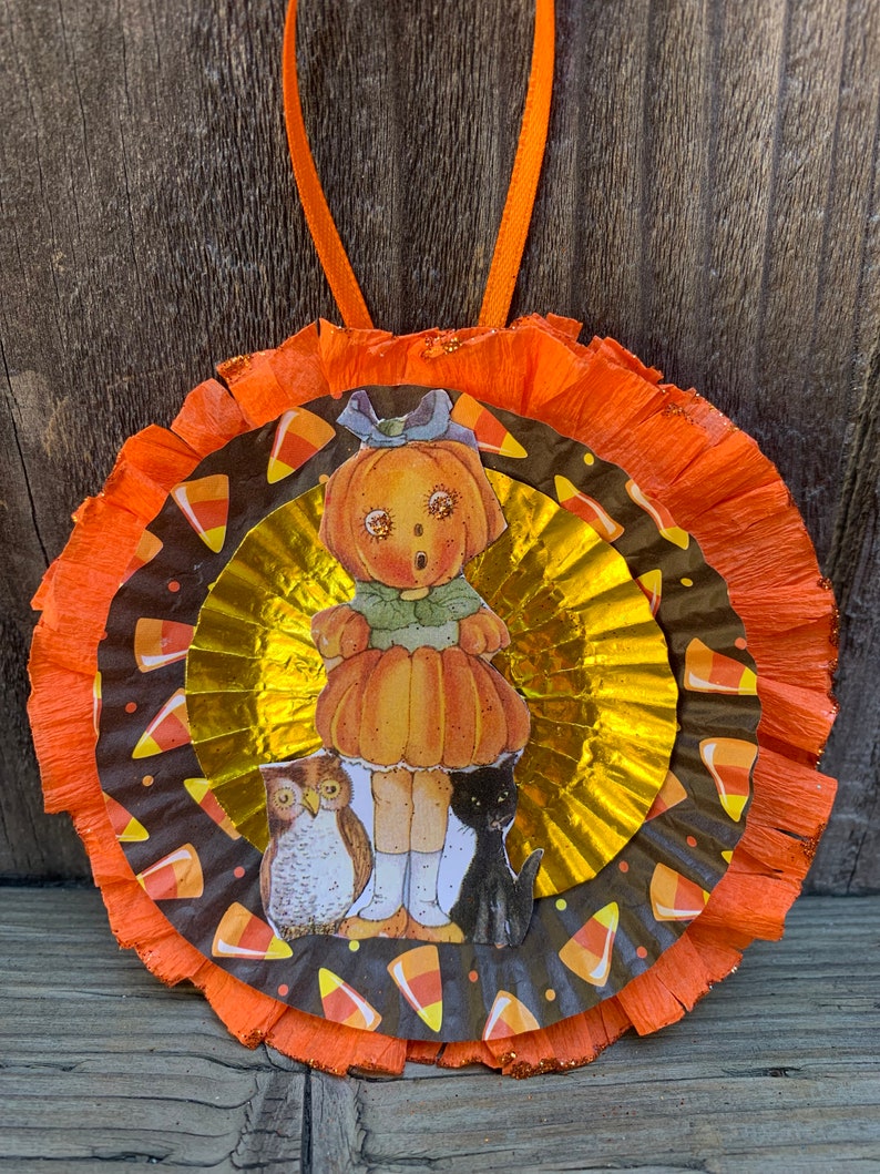 K&ouml;nnte beinhalten: Ein Halloween-Ornament mit einer K&uuml;rbiskopffigur, einer Eule und einer schwarzen Katze. Das Ornament hat Schichten aus orangefarbenem und goldenem Papier mit Zuckermais-Akzenten. Ein orangefarbenes Band ist zum Aufh&auml;ngen befestigt.