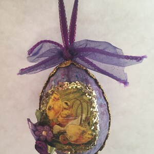 May include: Purple Easter egg decoration with a hanging ribbon. The egg has a decoupage image of yellow chicks and a floral decoration. The edges are trimmed with gold glitter.