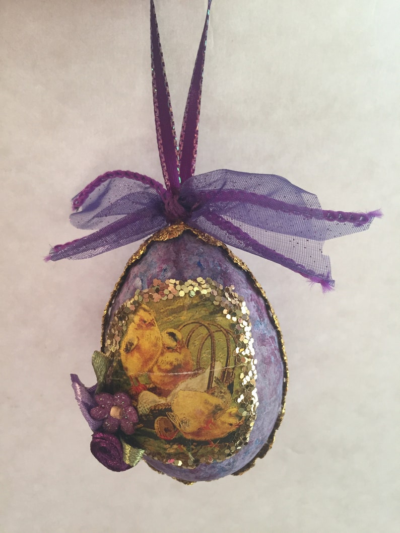 May include: Purple Easter egg decoration with a hanging ribbon. The egg has a decoupage image of yellow chicks and a floral decoration. The edges are trimmed with gold glitter.