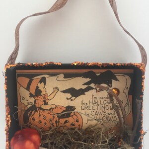 Pu&ograve; includere: Una scatola decorativa a tema Halloween con un'illustrazione vintage di un bambino e zucche. La scatola &egrave; nera con finiture glitterate arancioni e un manico in nastro marrone. L'illustrazione include il testo "I'm sending this Halloween greeting to be-CAWS well just be-CAWS."