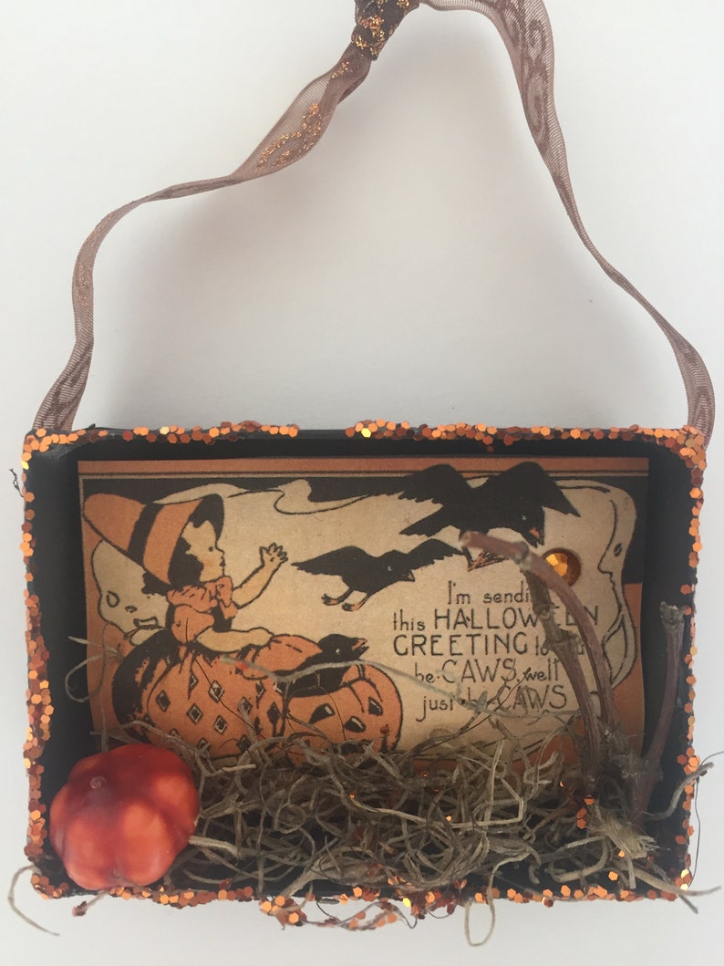 Pu&ograve; includere: Una scatola decorativa a tema Halloween con un'illustrazione vintage di un bambino e zucche. La scatola &egrave; nera con finiture glitterate arancioni e un manico in nastro marrone. L'illustrazione include il testo "I'm sending this Halloween greeting to be-CAWS well just be-CAWS."