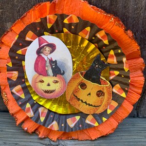 K&ouml;nnte beinhalten: Mehrschichtige Halloween-Dekoration mit orangefarbenen und braunen Papierkreisen. Das Design zeigt Vintage-Illustrationen eines Kindes mit Hexenhut, einer schwarzen Katze und K&uuml;rbislaternen. Die &auml;u&szlig;ere Schicht hat einen glitzernden Rand und ein orangefarbenes Band zum Aufh&auml;ngen.