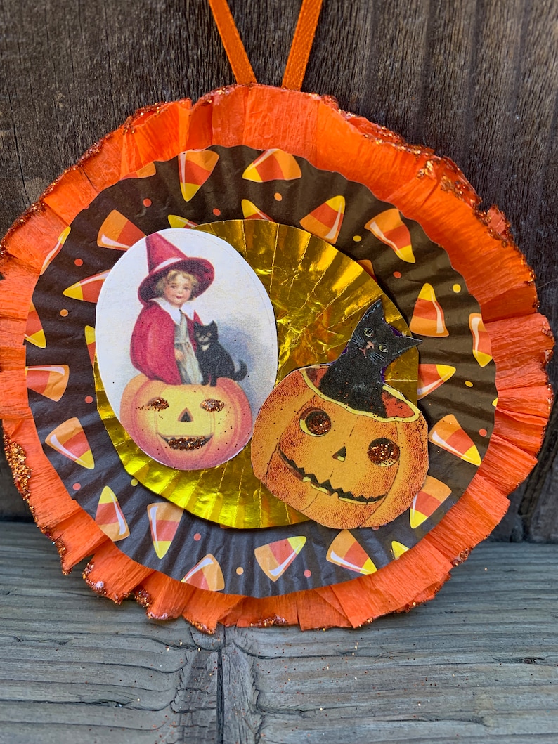 K&ouml;nnte beinhalten: Mehrschichtige Halloween-Dekoration mit orangefarbenen und braunen Papierkreisen. Das Design zeigt Vintage-Illustrationen eines Kindes mit Hexenhut, einer schwarzen Katze und K&uuml;rbislaternen. Die &auml;u&szlig;ere Schicht hat einen glitzernden Rand und ein orangefarbenes Band zum Aufh&auml;ngen.