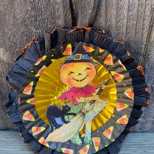 K&ouml;nnte beinhalten: Ein Halloween-Ornament mit einem l&auml;chelnden K&uuml;rbis-Charakter mit Hexenhut, der einen Besen h&auml;lt, mit einer schwarzen Katze. Das Ornament hat ein schwarz-goldenes, mehrschichtiges Design mit Zuckermais-Akzenten und einem schwarzen Band zum Aufh&auml;ngen.