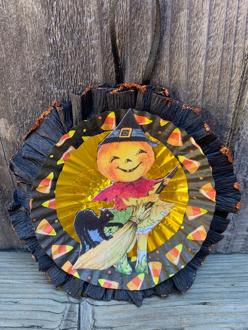K&ouml;nnte beinhalten: Ein Halloween-Ornament mit einem l&auml;chelnden K&uuml;rbis-Charakter mit Hexenhut, der einen Besen h&auml;lt, mit einer schwarzen Katze. Das Ornament hat ein schwarz-goldenes, mehrschichtiges Design mit Zuckermais-Akzenten und einem schwarzen Band zum Aufh&auml;ngen.