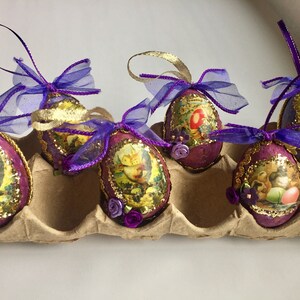 May include: Six decorative Easter egg ornaments. Each egg is oval-shaped, with a purple base, gold trim, and a vintage-style image. They are adorned with purple ribbons and small floral accents, resting in a cardboard egg carton.