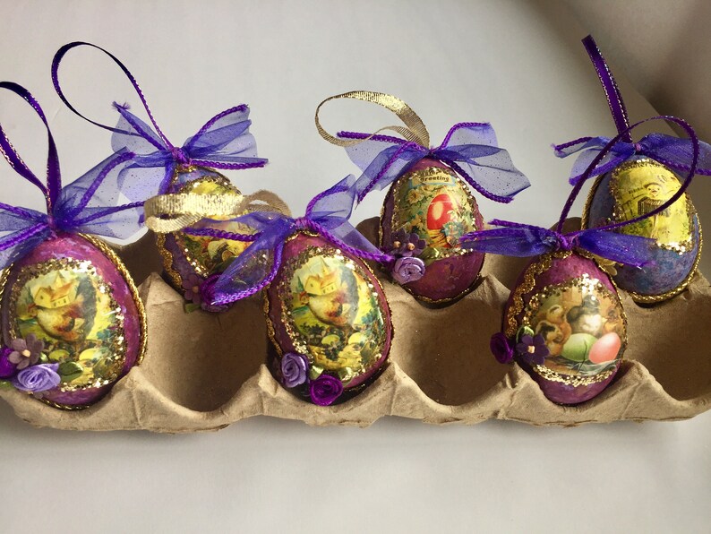 May include: Six decorative Easter egg ornaments. Each egg is oval-shaped, with a purple base, gold trim, and a vintage-style image. They are adorned with purple ribbons and small floral accents, resting in a cardboard egg carton.