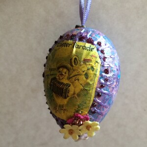 May include: Easter egg ornament with a textured purple and blue surface. The egg features a vintage image of a chick playing an accordion, with the words "Easter Parade". It is decorated with small flowers and a purple ribbon for hanging.