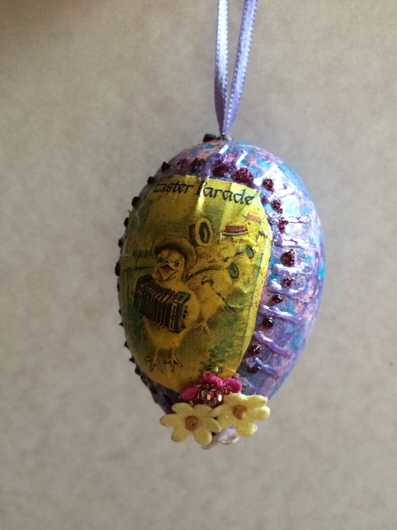 May include: Easter egg ornament with a textured purple and blue surface. The egg features a vintage image of a chick playing an accordion, with the words "Easter Parade". It is decorated with small flowers and a purple ribbon for hanging.