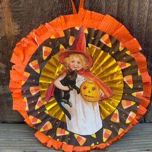 K&ouml;nnte beinhalten: Runde Halloween-Dekoration mit einer Vintage-Illustration eines Kindes mit Hexenhut, das eine schwarze Katze und einen K&uuml;rbis h&auml;lt. Die Dekoration hat Schichten aus orangefarbenem, goldenem und schwarzem Papier mit S&uuml;&szlig;igkeiten-Mais-Mustern.
