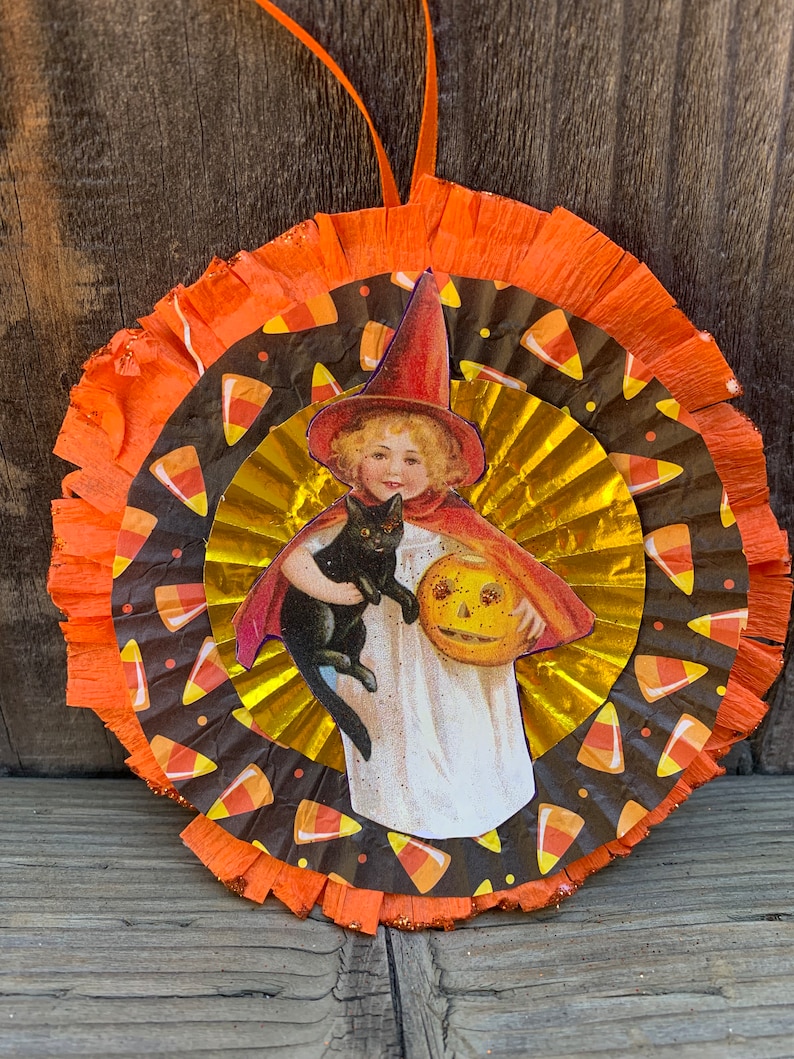 K&ouml;nnte beinhalten: Runde Halloween-Dekoration mit einer Vintage-Illustration eines Kindes mit Hexenhut, das eine schwarze Katze und einen K&uuml;rbis h&auml;lt. Die Dekoration hat Schichten aus orangefarbenem, goldenem und schwarzem Papier mit S&uuml;&szlig;igkeiten-Mais-Mustern.