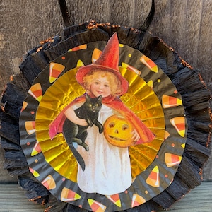 K&ouml;nnte beinhalten: Vintage Halloween-Dekoration mit einem zentralen Bild eines Kindes mit Hexenhut und Umhang, das eine schwarze Katze und einen K&uuml;rbis h&auml;lt. Das Bild befindet sich auf einer gold-schwarzen Papierrosette mit Candy-Corn-Akzenten und einem schwarzen Band zum Aufh&auml;ngen.