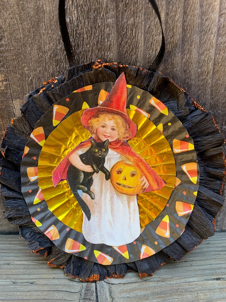 K&ouml;nnte beinhalten: Vintage Halloween-Dekoration mit einem zentralen Bild eines Kindes mit Hexenhut und Umhang, das eine schwarze Katze und einen K&uuml;rbis h&auml;lt. Das Bild befindet sich auf einer gold-schwarzen Papierrosette mit Candy-Corn-Akzenten und einem schwarzen Band zum Aufh&auml;ngen.