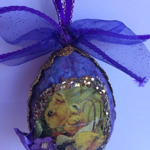 May include: A handcrafted Easter egg decoration. The egg is a deep purple hue, edged with gold, and features a picture of yellow chicks. It is embellished with a purple ribbon, bow, and a small purple flower.