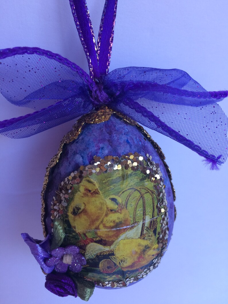 May include: A handcrafted Easter egg decoration. The egg is a deep purple hue, edged with gold, and features a picture of yellow chicks. It is embellished with a purple ribbon, bow, and a small purple flower.
