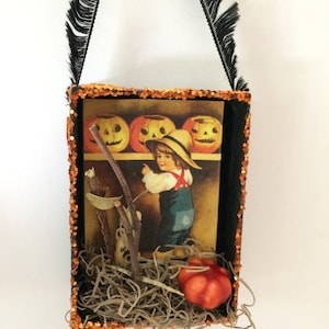Pu&ograve; includere: Una scatola ombra a tema Halloween con cornice nera e bordi glitterati arancioni. All'interno, un'immagine vintage di un bambino con zucche e una piccola zucca arancione. La scatola ha un manico in nastro nero con frange.