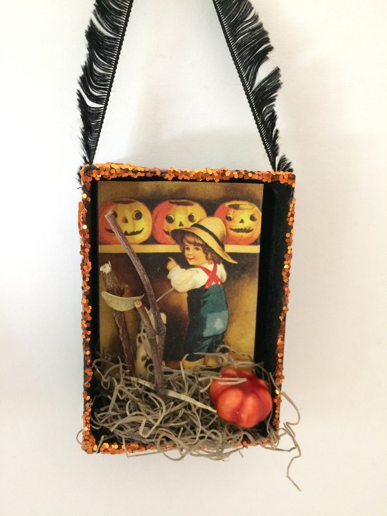 Pu&ograve; includere: Una scatola ombra a tema Halloween con cornice nera e bordi glitterati arancioni. All'interno, un'immagine vintage di un bambino con zucche e una piccola zucca arancione. La scatola ha un manico in nastro nero con frange.