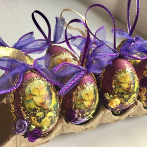 May include: Easter egg decorations in a cardboard egg box. The eggs are a deep purple colour with gold glitter trim and a picture of a landscape. Each egg has a purple and gold ribbon bow and small floral accents.
