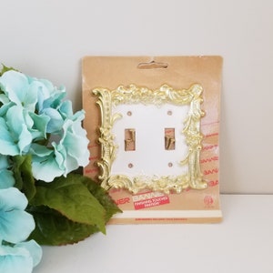 May include: A white double light switch plate with a gold ornate frame. The frame has a floral design and is packaged on a brown cardboard backing with the text "Banner Finishing Touches Finitiion".