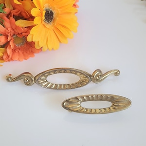 May include: Two gold-toned metal drawer pulls with ornate designs. One pull is oval-shaped with a decorative edge and a curved arm. The other pull is a simple oval shape.