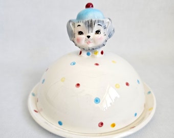 May include: A white ceramic butter dish with a lid featuring a cartoon dog wearing a blue hat. The dish is decorated with colorful polka dots.