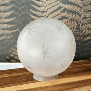 May include: Frosted glass globe with etched starburst patterns. The round light fixture sits on a small base. The background features a fern pattern.