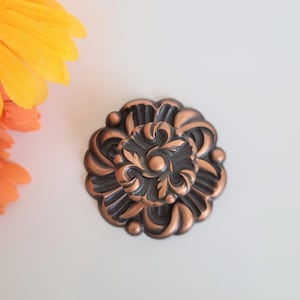 May include: A decorative copper-coloured metal embellishment with a floral design. The embellishment has a dark brown centre with a raised, circular, copper-coloured centre.