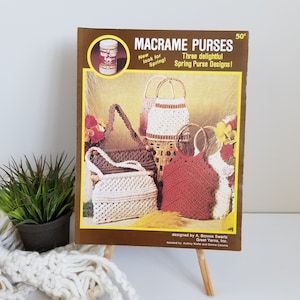 May include: A vintage craft book with a brown cover featuring a photo of three macrame purses. The title of the book is "Macrame Purses" and the text "Three delightful Spring Purse Designs!" is printed below the title. The book is designed by A. Bonnie Swartz, Great Yarns, Inc. and assisted by Audrey Kiefer and Donna Caronia.