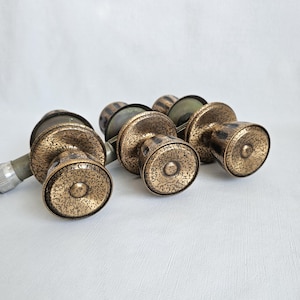 May include: A collection of antique brass door knobs with a textured, speckled pattern. The knobs feature a circular base and a cylindrical body. The metal has a tarnished, aged look, indicating they are vintage or vintage-style.
