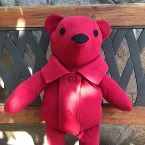 May include: A red teddy bear wearing a red collared shirt with buttons. The bear is sitting on a wooden bench.