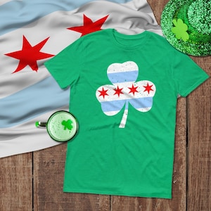 May include: A green t-shirt featuring a shamrock design with the Chicago city flag incorporated. The shamrock has white, light blue, and red star accents. A green beer, a green hat, and a Chicago flag are also in the image.