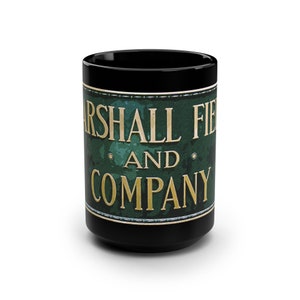 Op de afbeelding: Zwarte keramische mok met een groen en gouden ontwerp. Het ontwerp bevat de tekst "Marshall Field and Company".