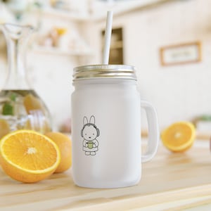 Può includere: Tazza bianca in barattolo Mason con coperchio e manico argentati, con un coniglietto dei cartoni animati con le cuffie e che tiene una tazza verde. Una cannuccia bianca è inserita nella tazza. La tazza è su una superficie di legno con fette d'arancia.