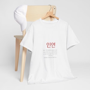 Può includere: T-shirt bianca con la parola coreana "인연" in rosso e una citazione in inglese. Un berretto da baseball bianco è appoggiato su una sedia di legno. La maglietta è appesa a un appendiabiti di legno.