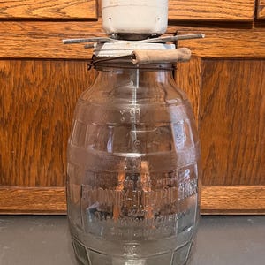 May include: An antique glass churn with a white electric motor on top. The churn has a wooden handle and metal components. The text on the glass reads "Dandy Electric Churn". The churn is on a gray surface.
