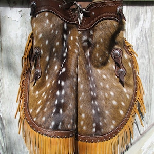 May include: A pair of brown and white spotted leather chaps with brown leather fringe and brown leather straps. The chaps have a brown leather trim and a brown leather buckle.
