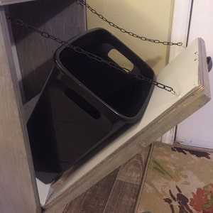 May include: A wooden cabinet with a hinged door and a black bin inside. The door is open and the bin is hanging from a chain.