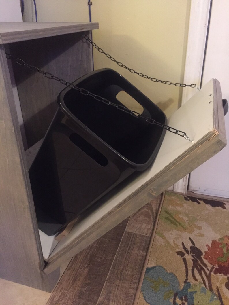 May include: A wooden cabinet with a hinged door and a black bin inside. The door is open and the bin is hanging from a chain.