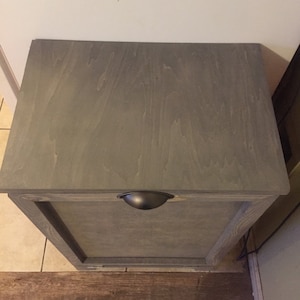 May include: A grey wooden cabinet with a single door and a black metal handle. The cabinet is sitting on a wood floor.