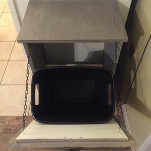 May include: A grey wooden cabinet with a hinged top and a black bin hanging from chains. The cabinet is open and the bin is visible.