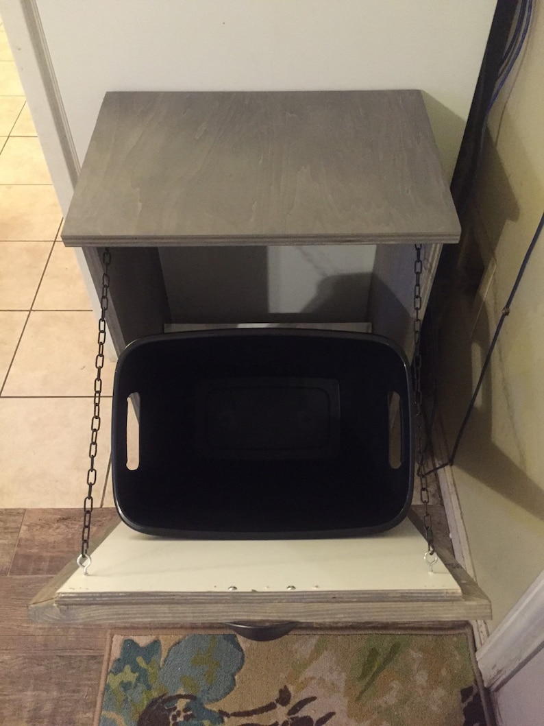 May include: A grey wooden cabinet with a hinged top and a black bin hanging from chains. The cabinet is open and the bin is visible.