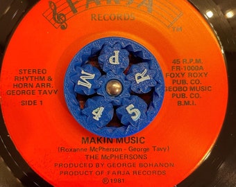 Könnte beinhalten: Eine rote Schallplatte mit dem Titel "Makin Music" von The McPhersons. Das Plattenlabel enthält außerdem den Text "Stereo Rhythm & Horn Arr. George Tavy, Side 1, Faria Records, 45 R.P.M. FR-1000A, Foxy Roxy Pub. Co., Geobo Music Pub. Co. B.M.I., ℗ 1981".
