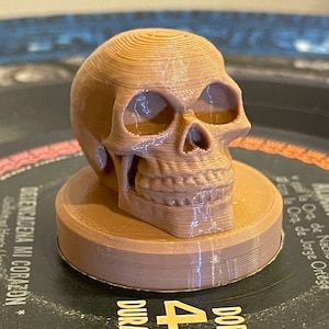 May include: A 3D printed brown skull figurine with a detailed design. The skull is sitting on a round base and is made of a layered material.