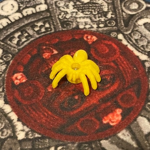 K&ouml;nnte beinhalten: Ein gelbes Plastik-Spinnenspielzeug mit acht Beinen und einem kleinen Loch in der Mitte. Die Spinne sitzt auf einer rot-schwarz gemusterten Oberfl&auml;che.