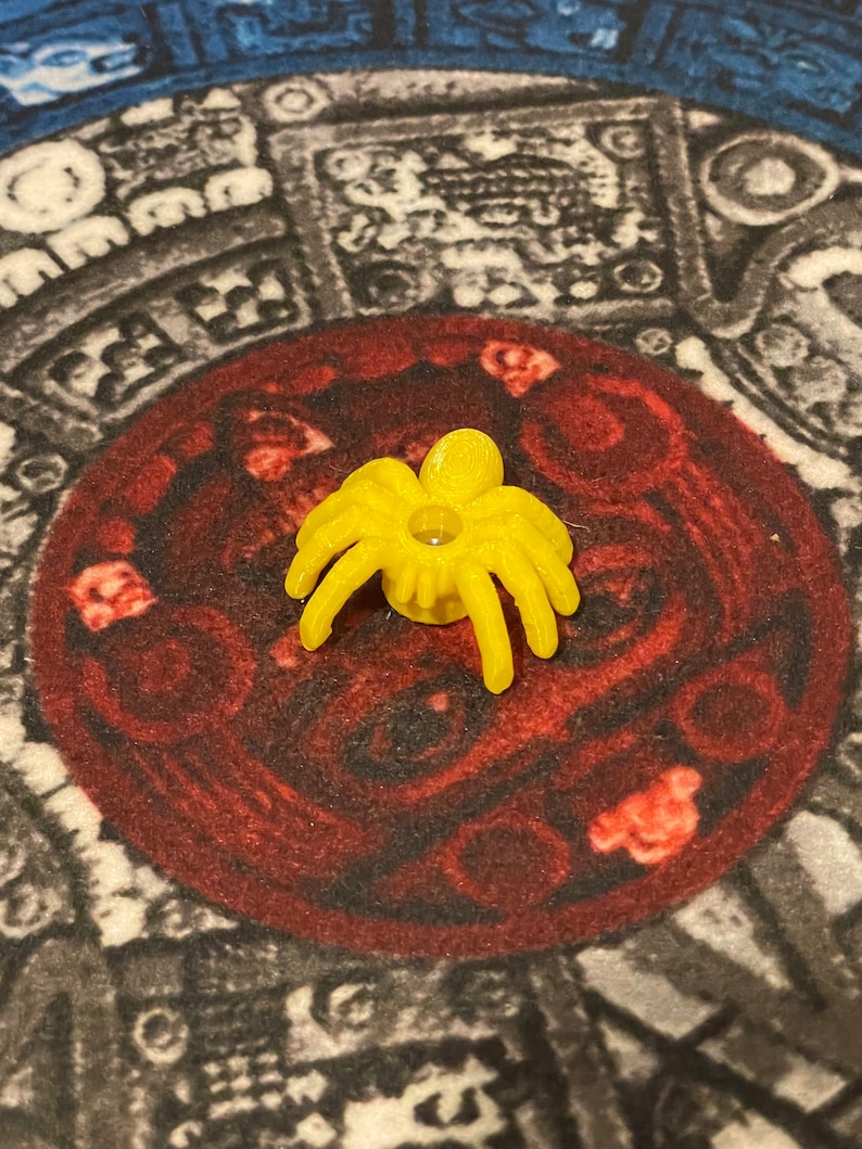 K&ouml;nnte beinhalten: Ein gelbes Plastik-Spinnenspielzeug mit acht Beinen und einem kleinen Loch in der Mitte. Die Spinne sitzt auf einer rot-schwarz gemusterten Oberfl&auml;che.